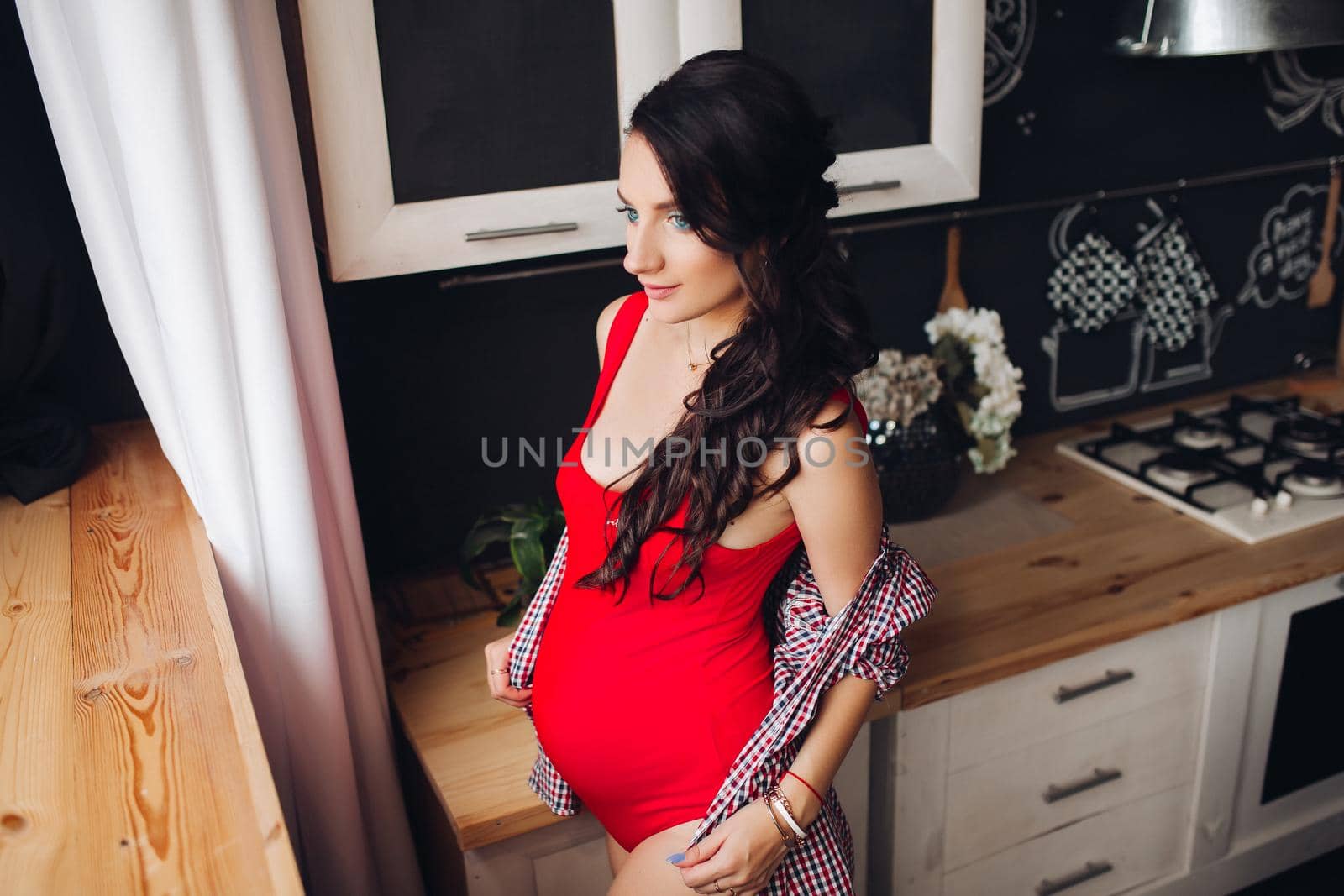 Adorable pregnant woman standing at kitchen and touching big belly with hands. Attractive brunette expecting little child and enjoying motherhood. Concept of love and pregnancy.