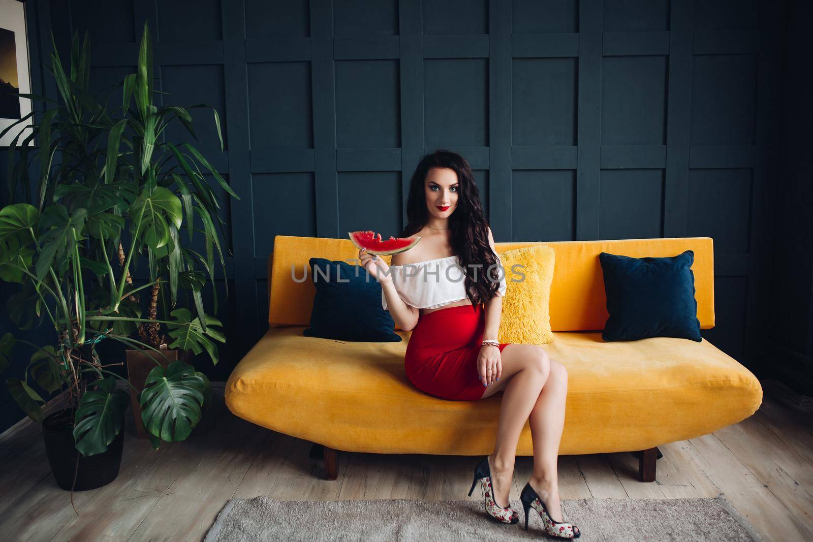 Stylish pregnant woman sitting on orange sofa, eating watermelon and posing. Attractive future mother in elegant clothes awaiting for little baby and relaxing. Concept of pregnancy and fashion.