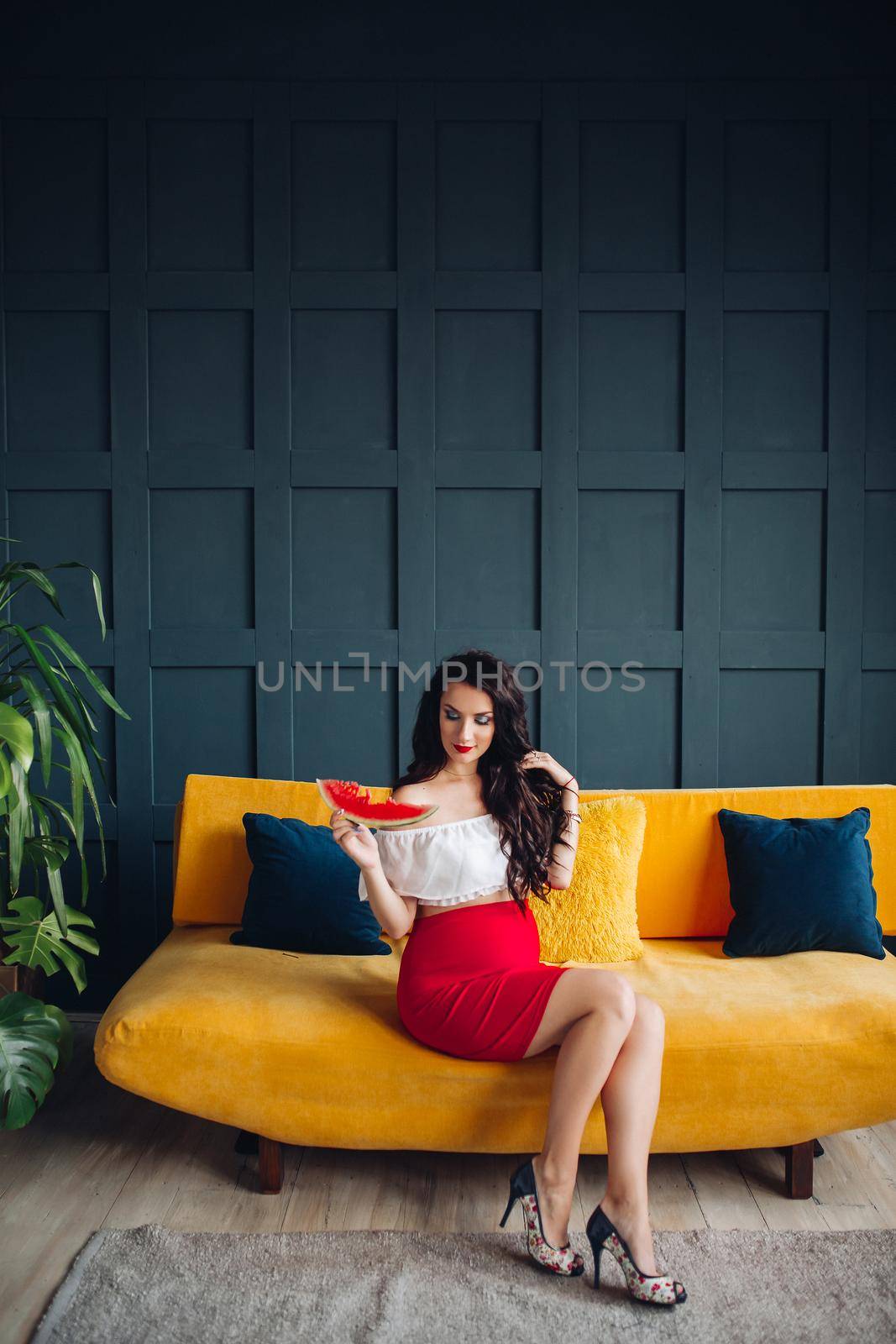 Stylish pregnant woman sitting on orange sofa, eating watermelon and posing. Attractive future mother in elegant clothes awaiting for little baby and relaxing. Concept of pregnancy and fashion.