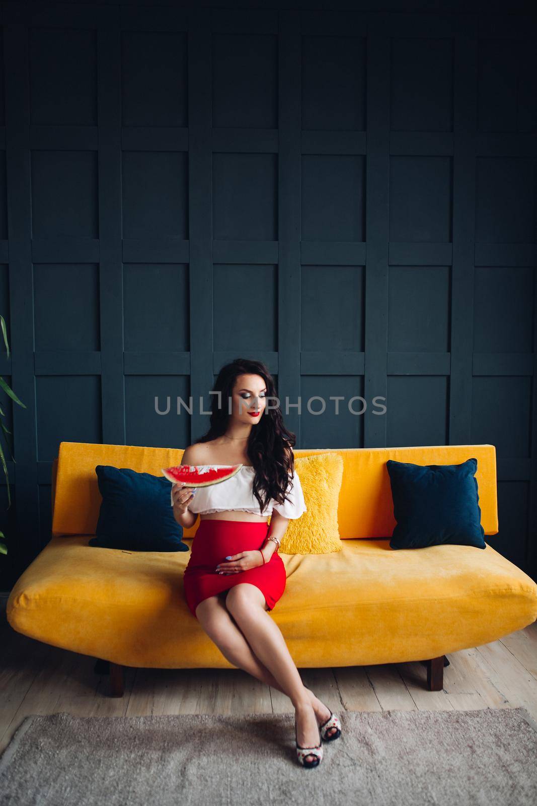 Stylish pregnant woman sitting on orange sofa and posing by StudioLucky