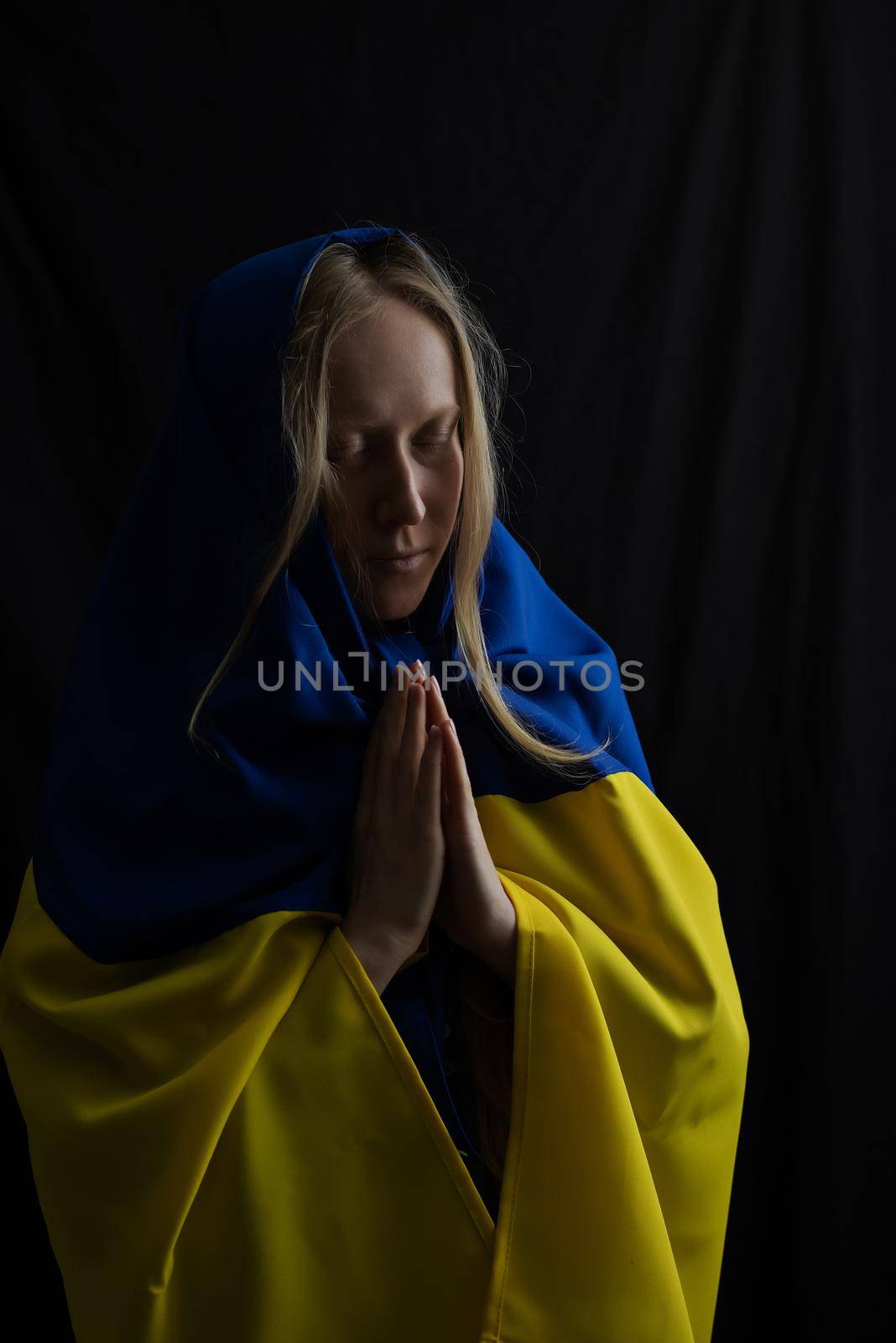 Ukrainian blonde woman prays with closed eyes for Ukraine during the war with the state flag on her head. Russia attacked Ukraine on February 24, 2022