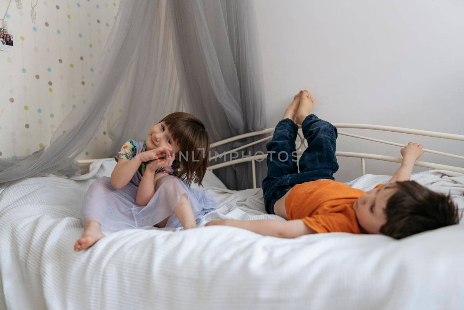 Two siblings happily playing together on the sofa bed covered with white blanket in the kids' room smiling into the camera. High quality photo