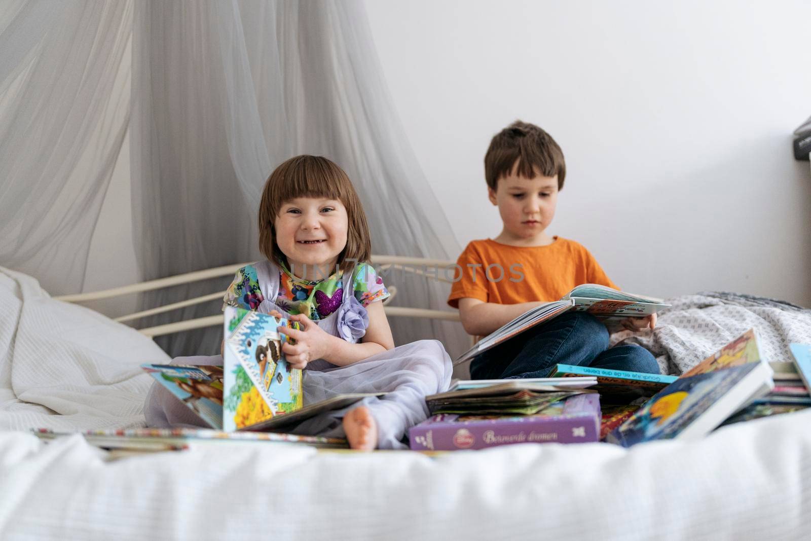 Two siblings reading books together on the sofa bed covered with white blanket in the kids' room, one of them smiling into the camera