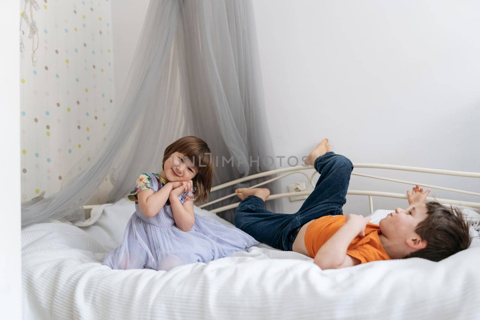 Two siblings happily playing together on the sofa bed covered with white blanket in the kids' room smiling into the camera. High quality photo