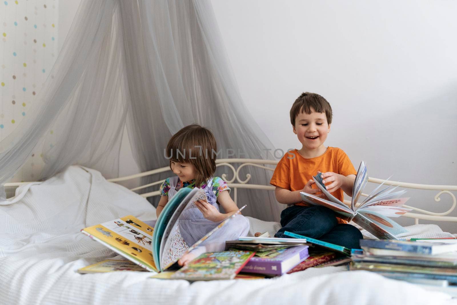 Two siblings reading books together on the sofa bed covered with white blanket in the kids' room