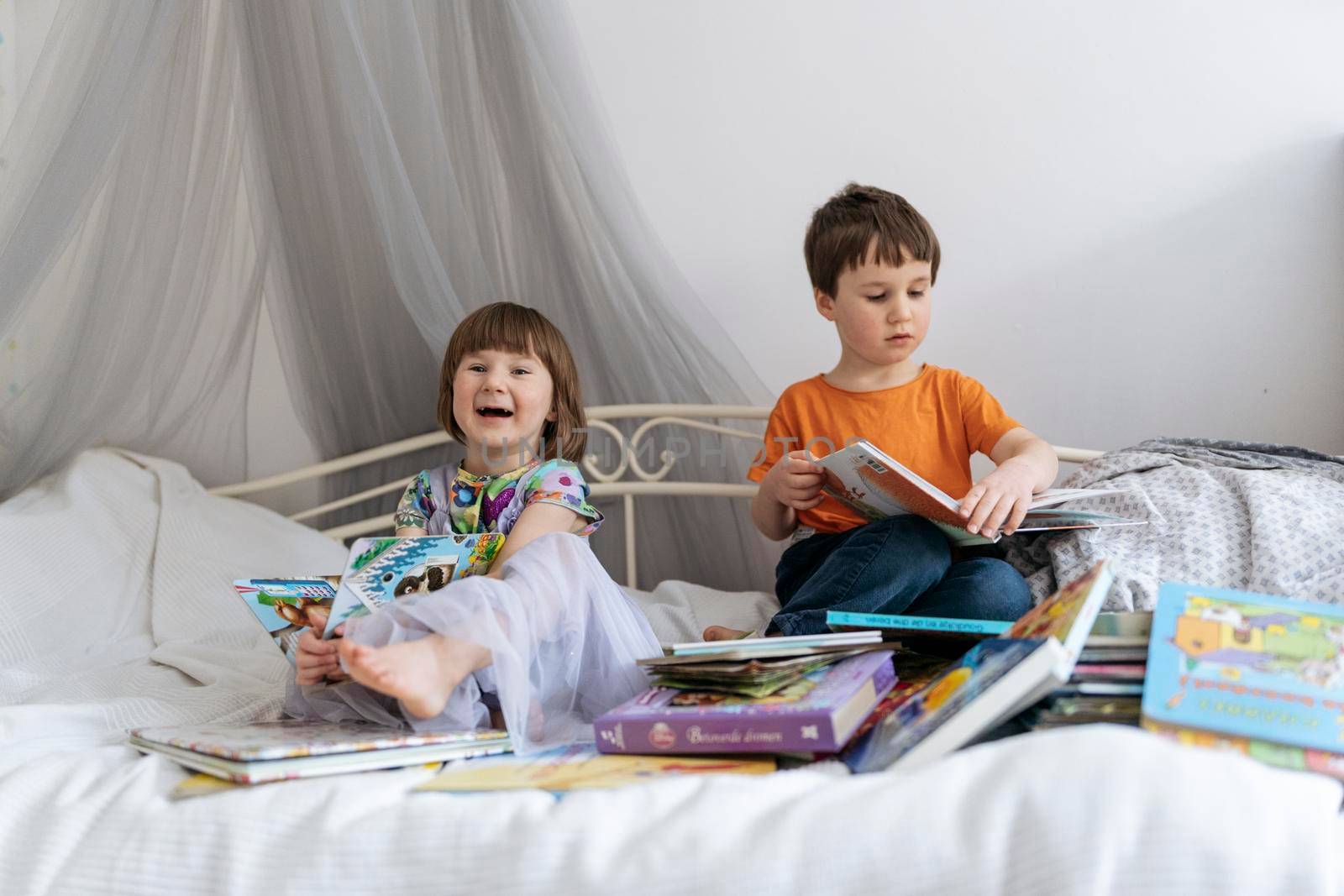 Two siblings reading books together on the sofa bed covered with white blanket in the kids' room, while both of them are smiling