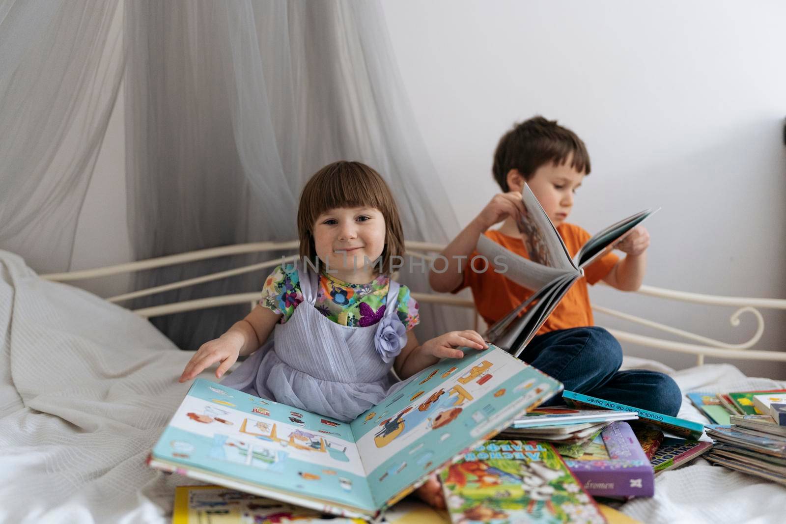 Two siblings reading books together on the sofa bed covered with white blanket in the kids' room, one of them laughing into the camera