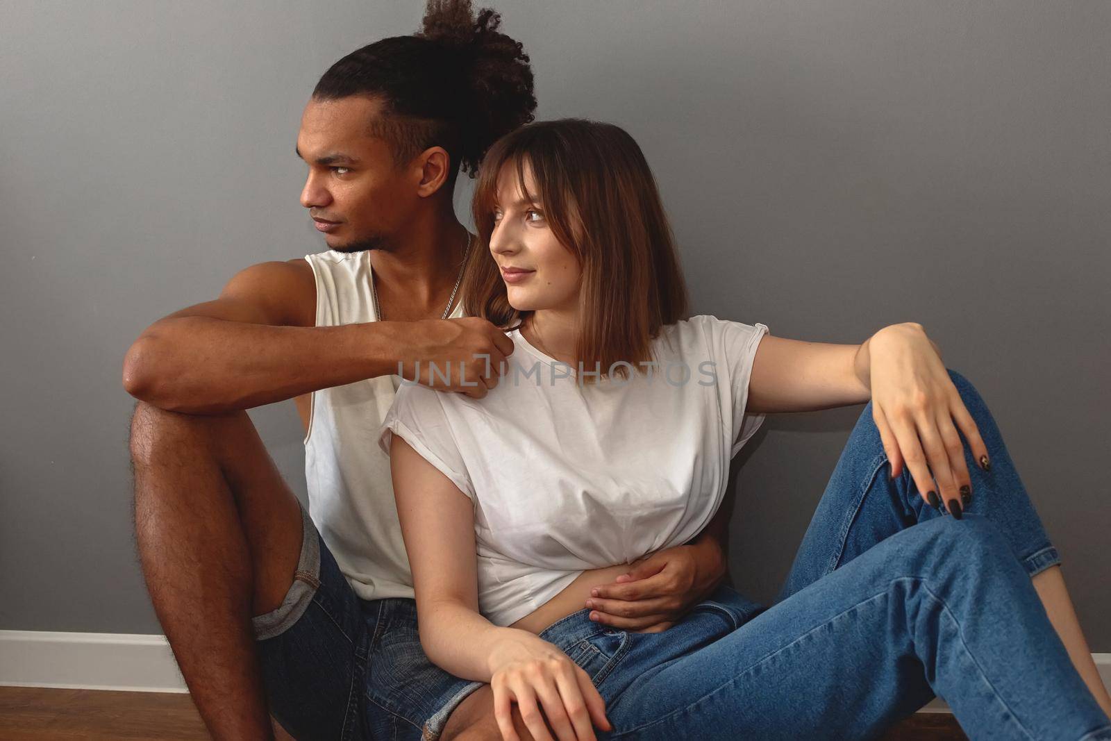 Multiracial family, stylish guy and girl in white t-shirts and blue jeans are sitting on the floor at home against a gray wall looking away.