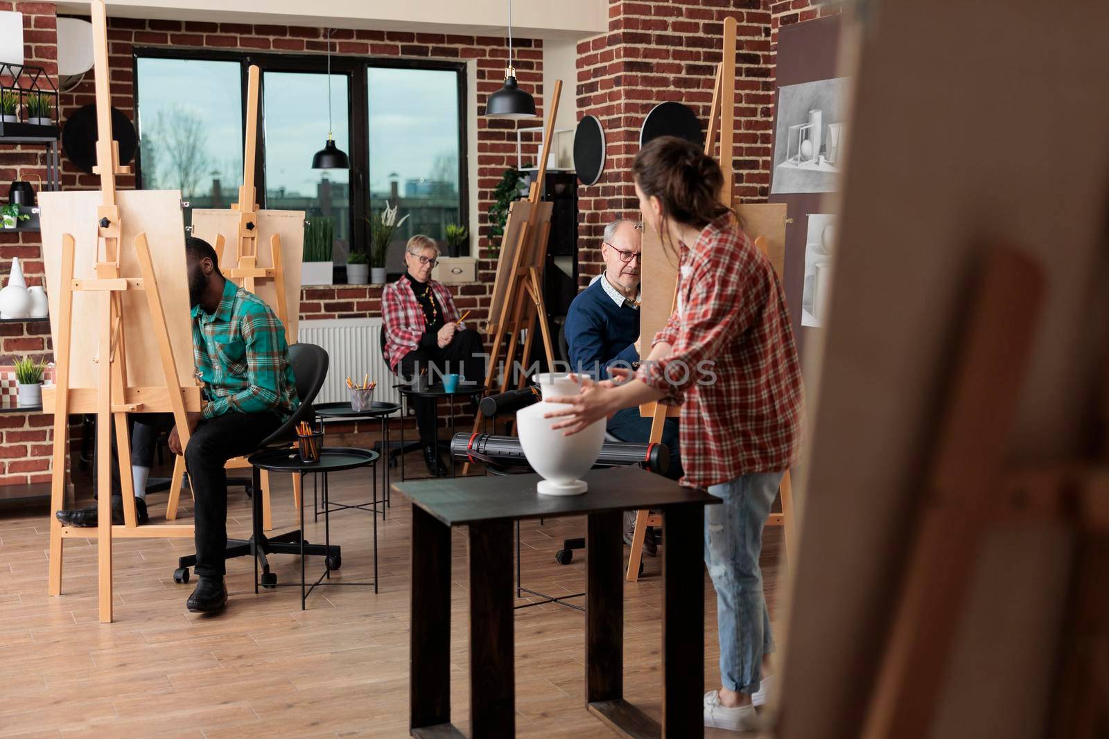 Young teacher showing inspiration for drawing course explaining illustration technique during art lesson in creativity studio. Multiethnic team sketching vase on painting canvas using graphic pencil