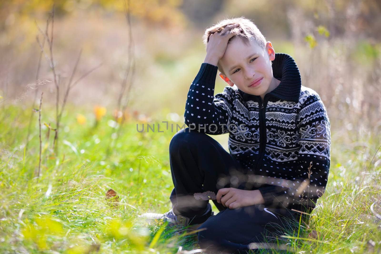 A handsome calm boy in a knitted sweater sits on the grass on the field.