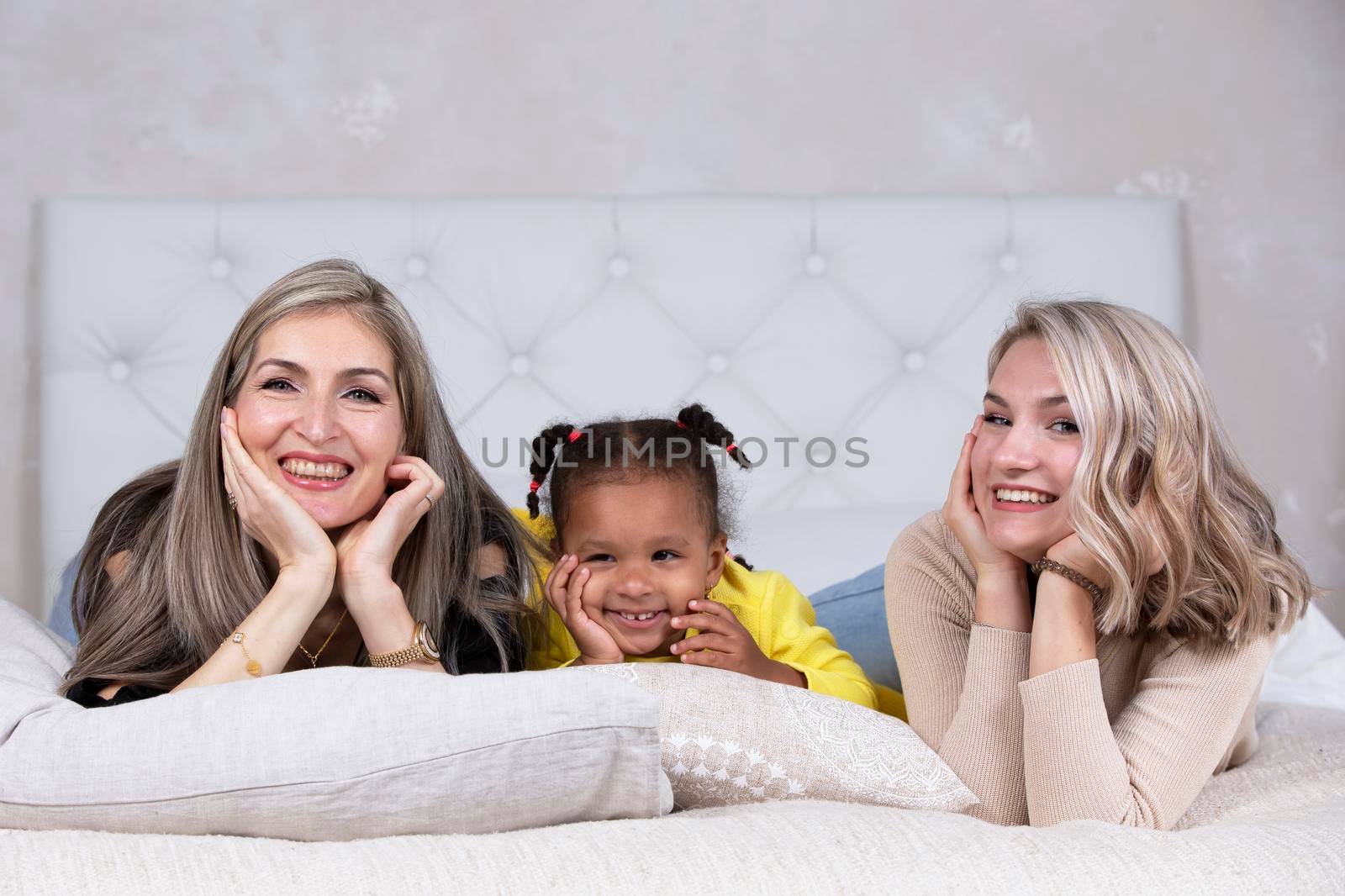 Family from different races. Mom grandmother light-skinned and a little African American girl lie on the sofa. by Sviatlana