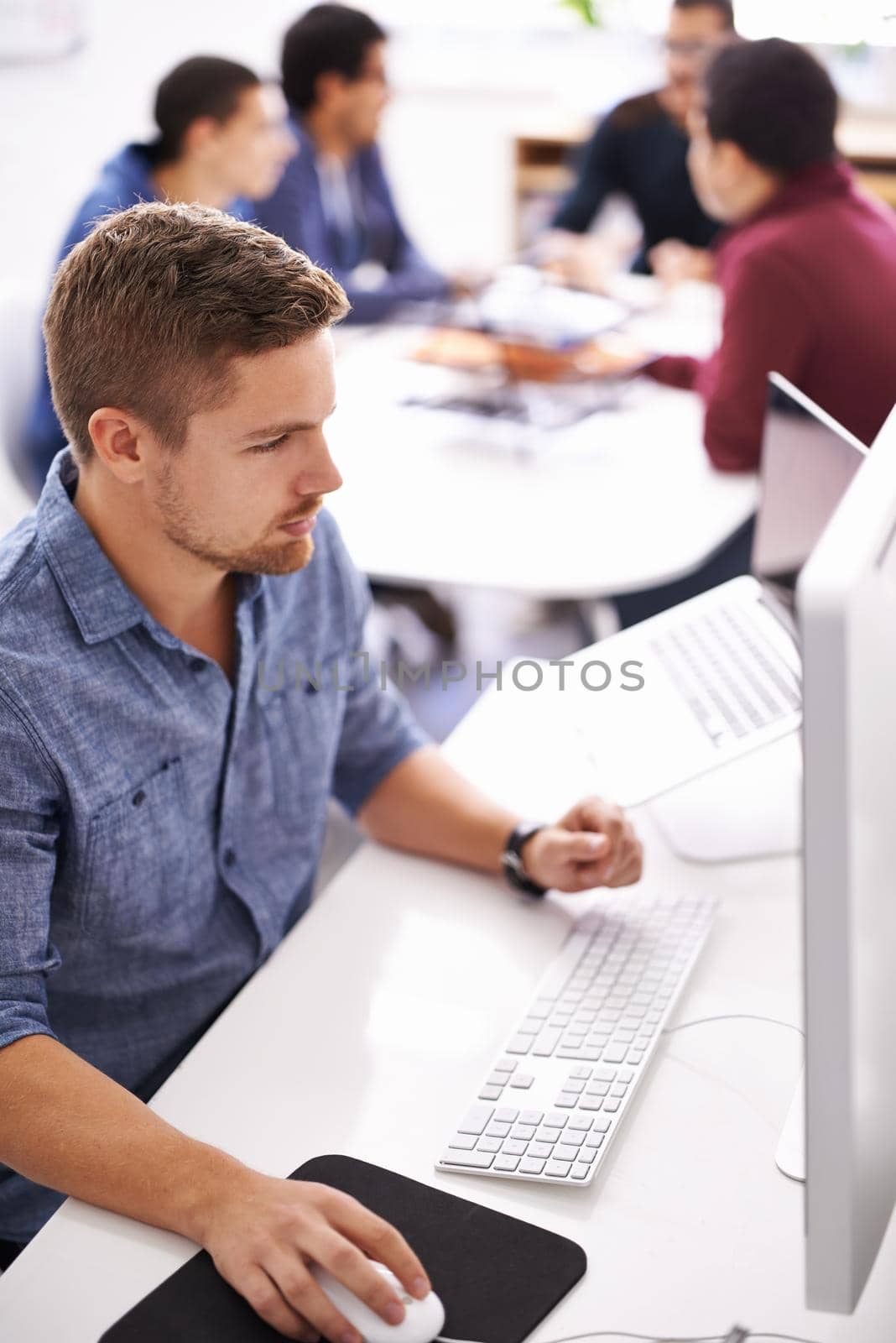 Shot of a young designer working at his computer with colleagues in the background.