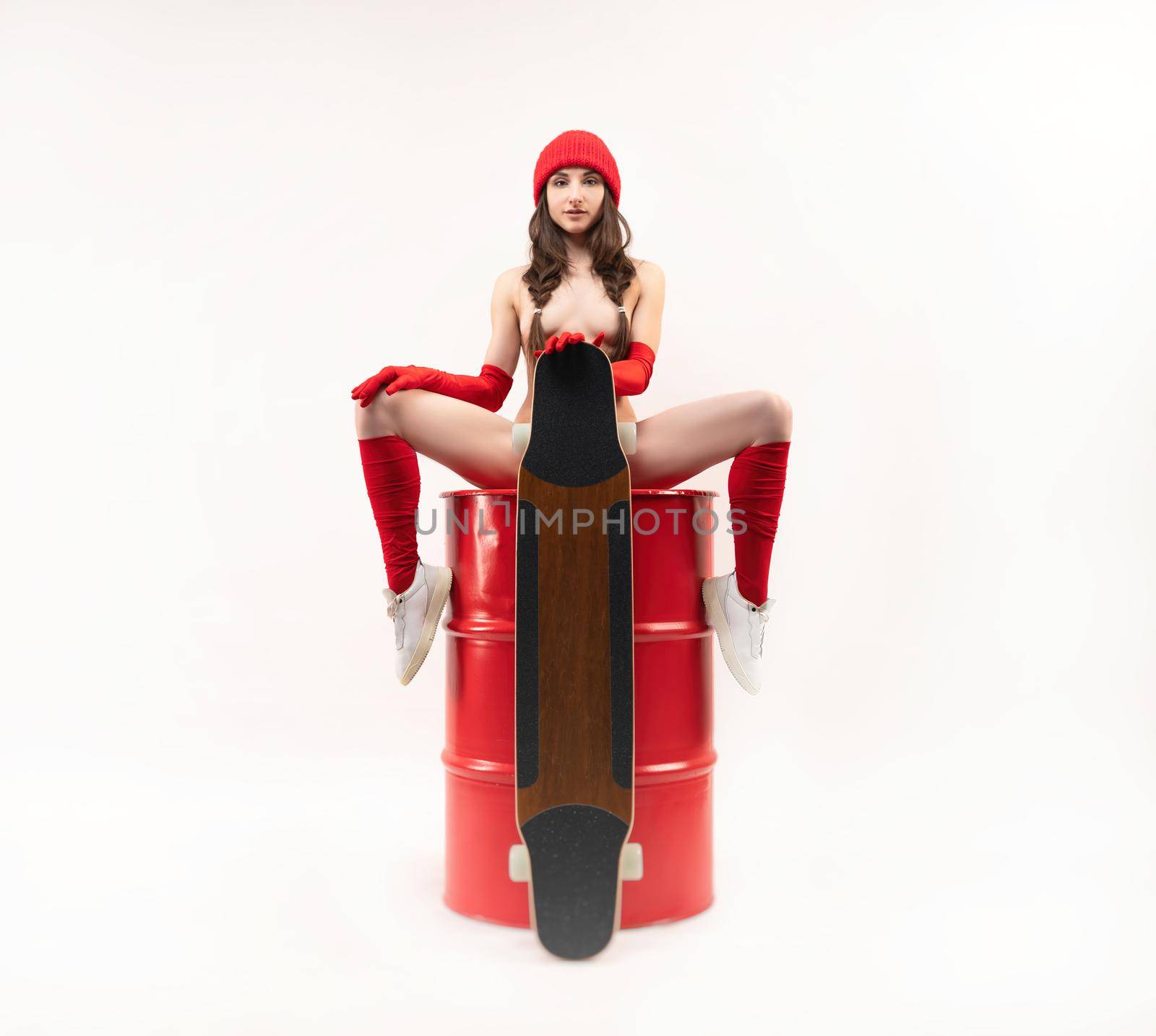 naked skater girl with a longboard in a red hat and socks sits sexy on a red barrel on a white background