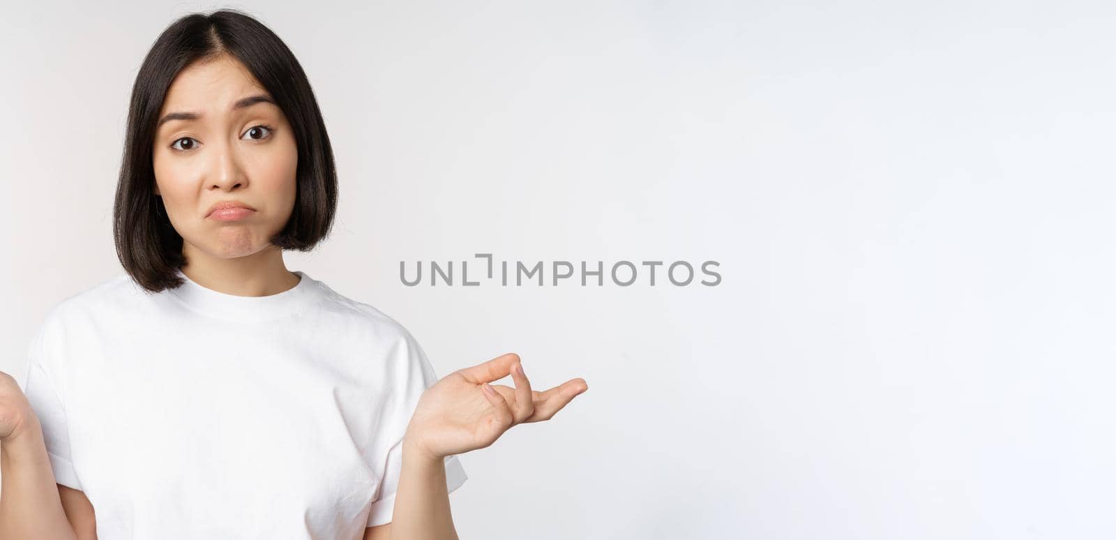 Portrait of confused asian woman shrugging shoulders, looking clueless, puzzled to say, standing over white background. Copy space