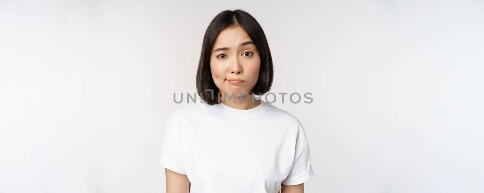 Image of young asian brunette woman in white t-shirt, smirk and look with doubt, hesitating, standing against white background. Copy space