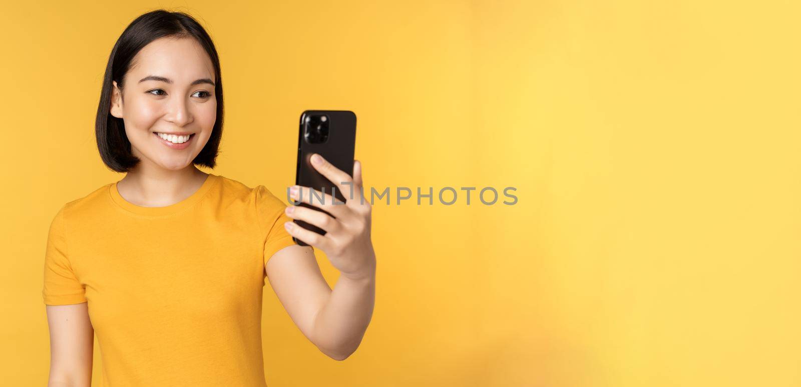 Image of happy, beautiful asian girl video chatting, talking on smartphone application, standing against yellow background by Benzoix