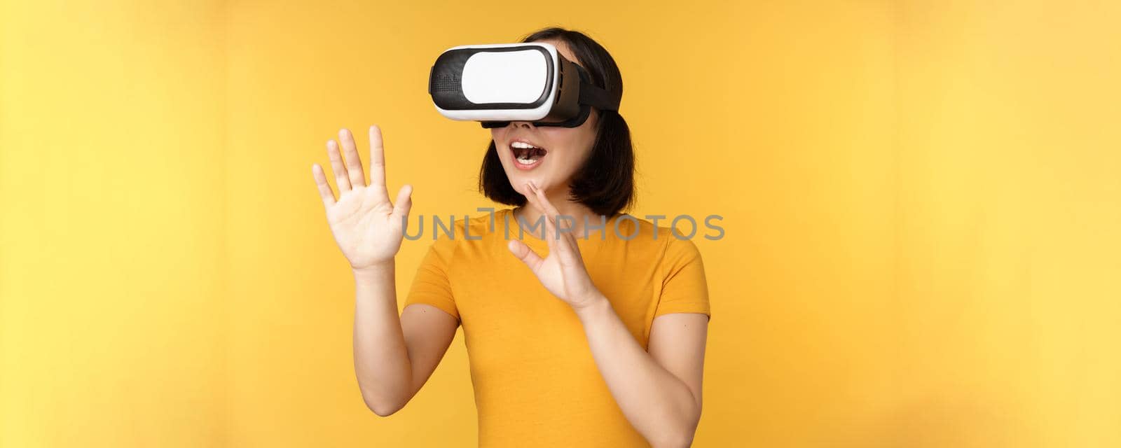 Girl in VR. Beautiful young asian woman, using virtual reality glasses and playing, chatting virtually, standing over yellow background.