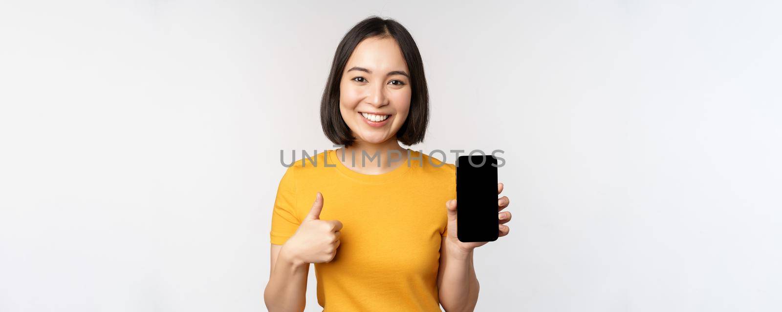 Technology and people concept. Smiling young woman showing thumb up and smartphone screen, mobile phone app interface, standing against white background by Benzoix
