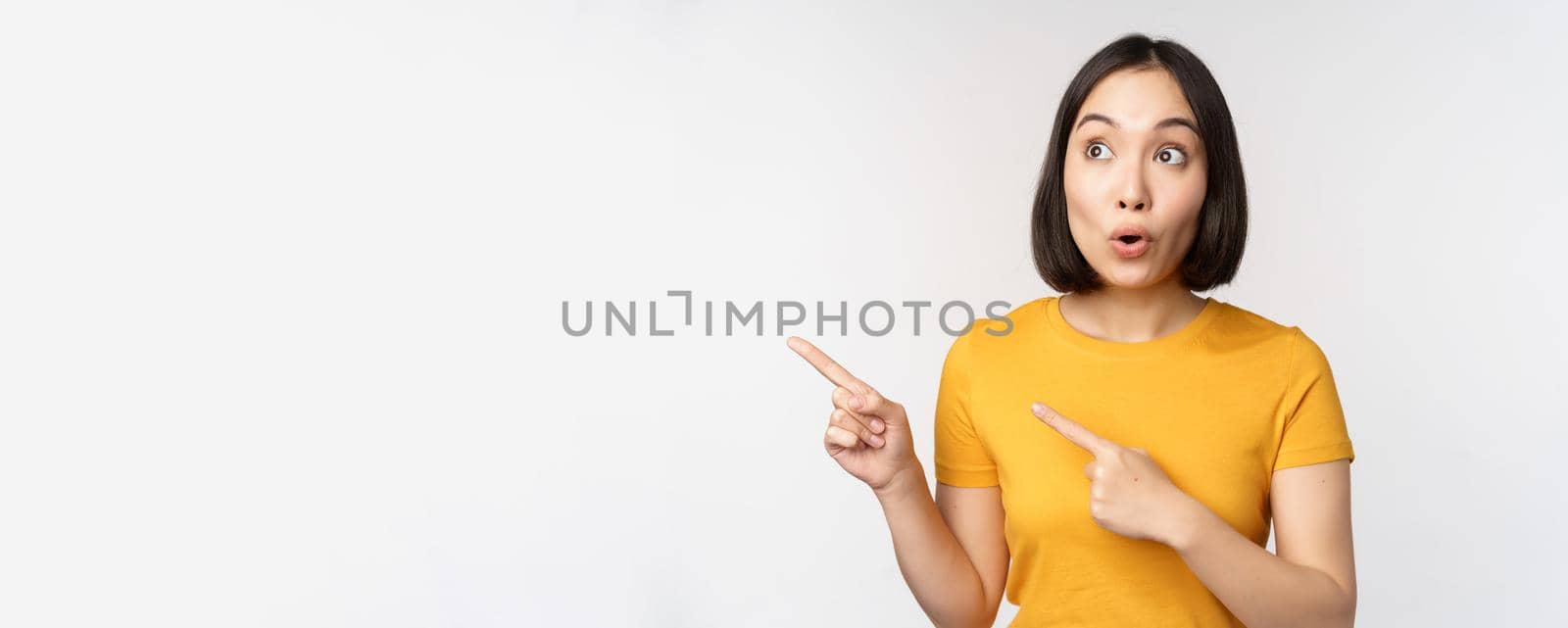 Surprised asian woman pointing and looking left at copy space, promo sale, showing advertisement with impressed face expression, standing over white backround.