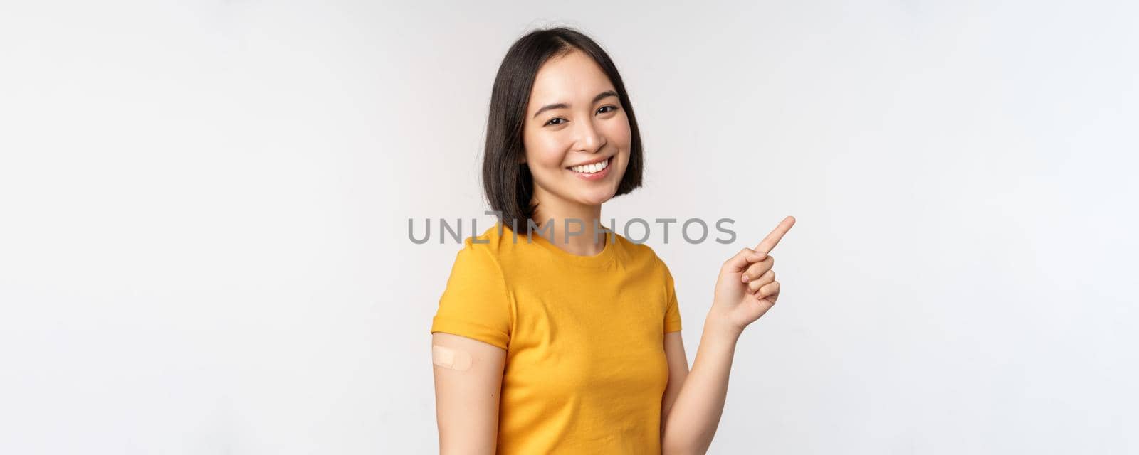Covid-19 and vaccination concept. Smiling asian girl with band aid on shoudler, pointing finger at banner, showing vaccine campaign, standing over white background.