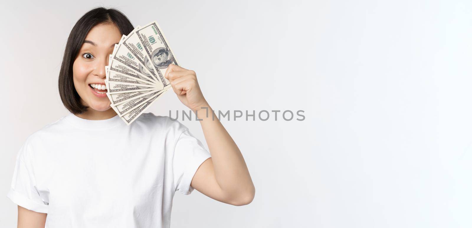 Portrait of smiling asian woman holding dollars money, concept of microcredit, finance and cash, standing over white background.