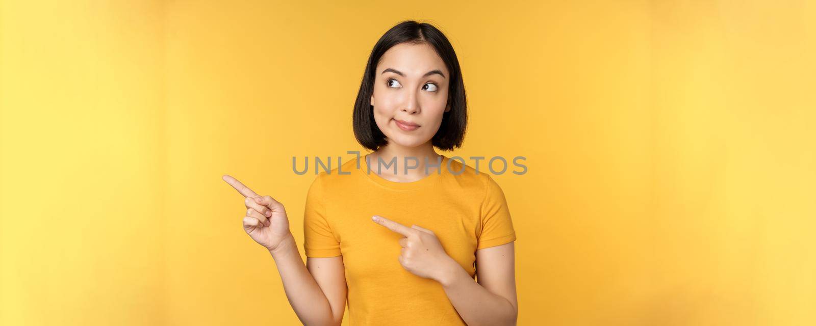 Portrait of intrigued asian woman, looking and pointing fingers left at advertisement, showing smth interesting, standing over yellow background by Benzoix