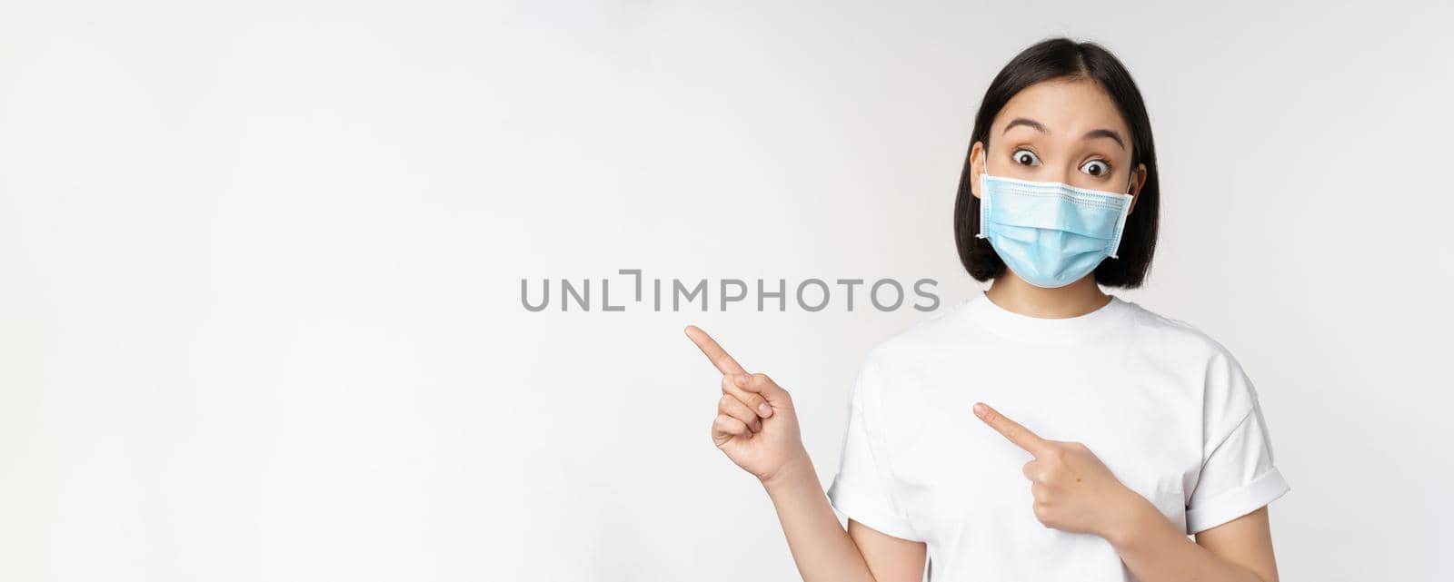 Surprised asian girl in medical mask, pointing fingers left, showing promo offer, raising eyebrows, amazed reaction, standing against white background.