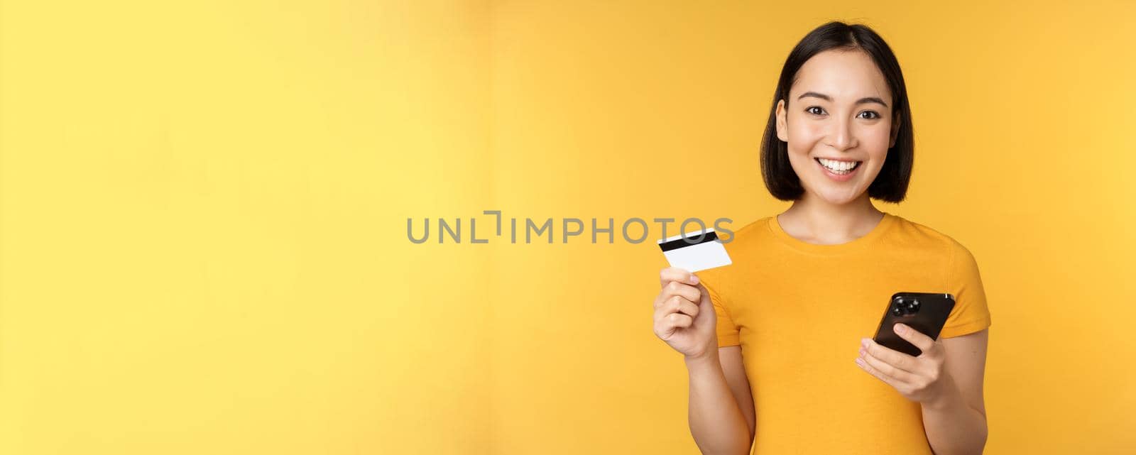 Online shopping. Cheerful asian girl holding credit card and smartphone, paying, order with mobile phone, standing over yellow background by Benzoix