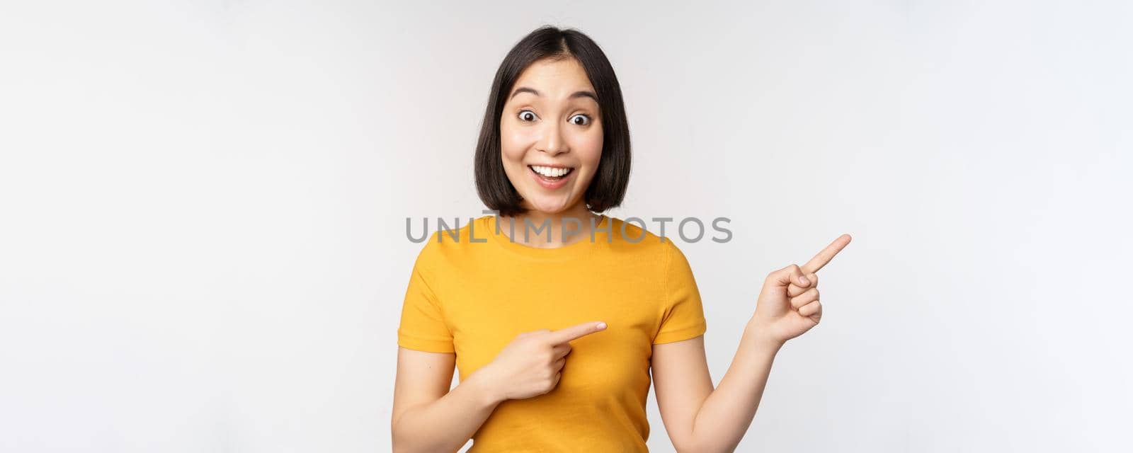 Amazed young asian woman, showing advertisement aside, pointing fingers right at promotion text, brand logo, standing happy against white background.