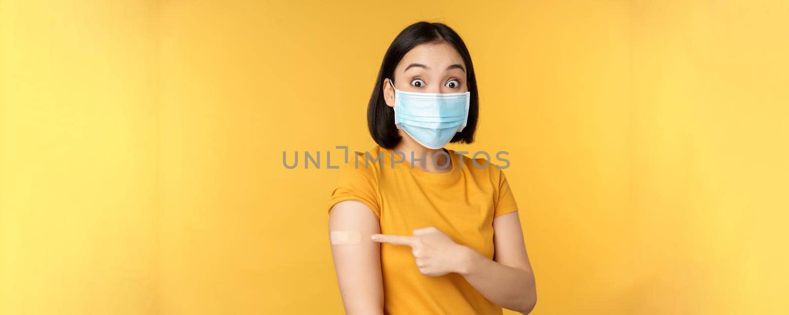 Vaccination from covid and health concept. Excited asian woman in medical face mask, pointing finger at shoulder with band aid, standing over yellow background.