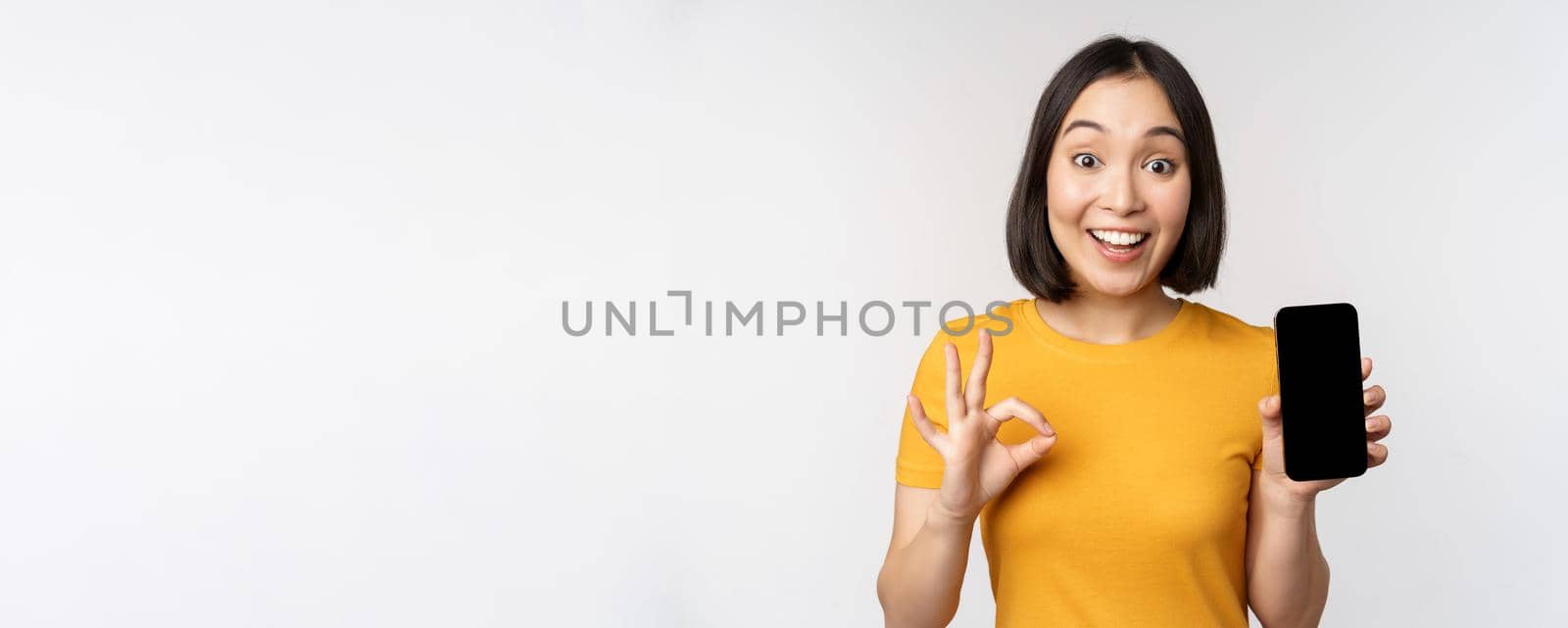 Excited asian girl showing mobile phone screen, okay sign, recommending smartphone app, standing in yellow tshirt over white background.
