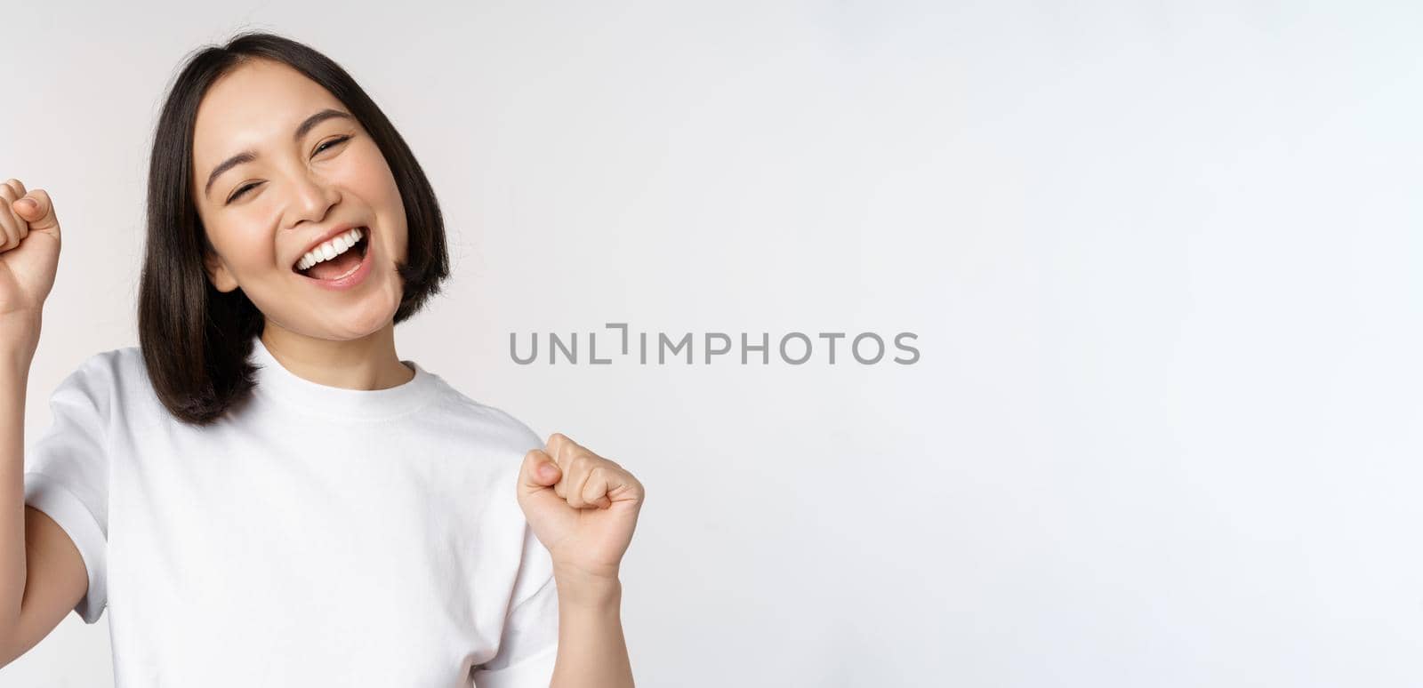 Dancing asian girl celebrating, feeling happy and upbeat, smiling broadly, standing over studio white background by Benzoix