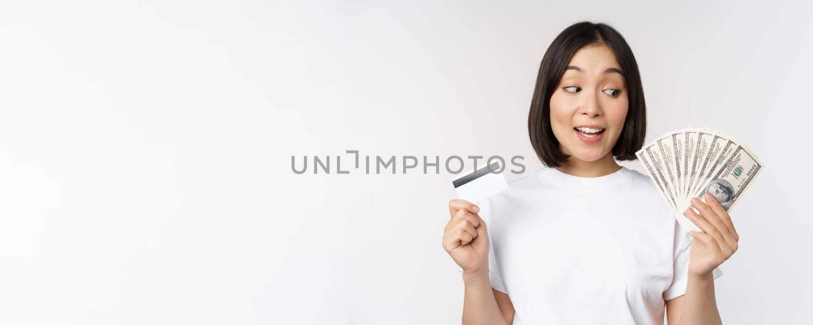 Portrait of asian woman holding money, dollars and credit card, looking impressed and amazed, standing in tshirt over white background by Benzoix