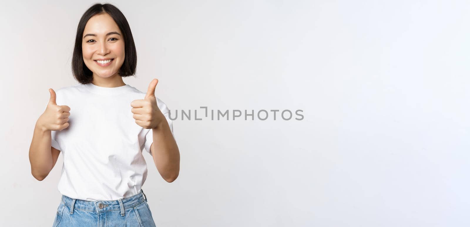 Happy beautiful korean girl, showing thumbs up in approval, smiling pleased, like smth, recommending, standing over white background.