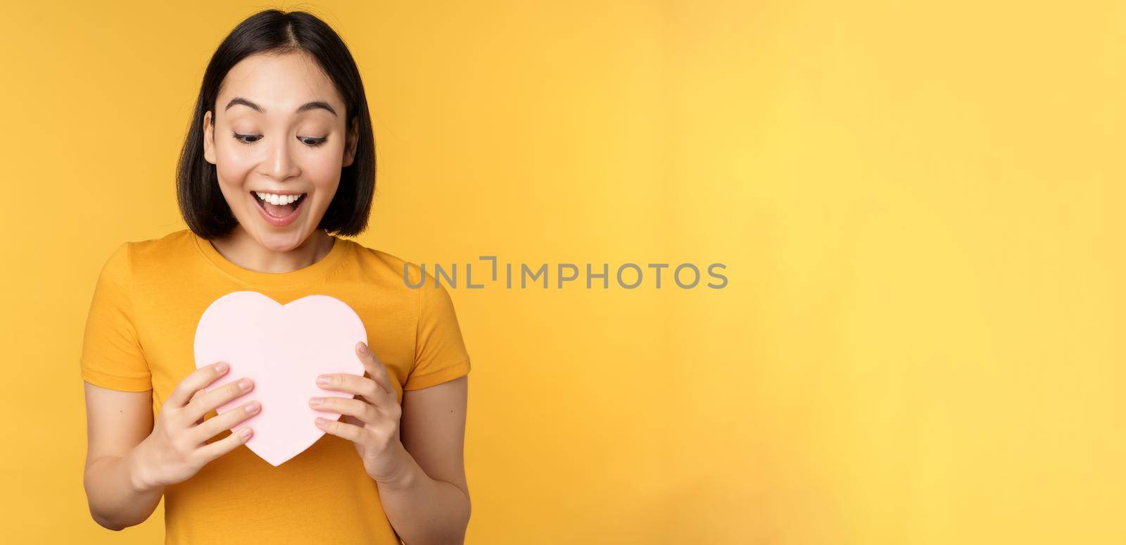 Romance and valentines day. Happy beautiful asian woman holding big heart card and smiling, standing over yellow background by Benzoix