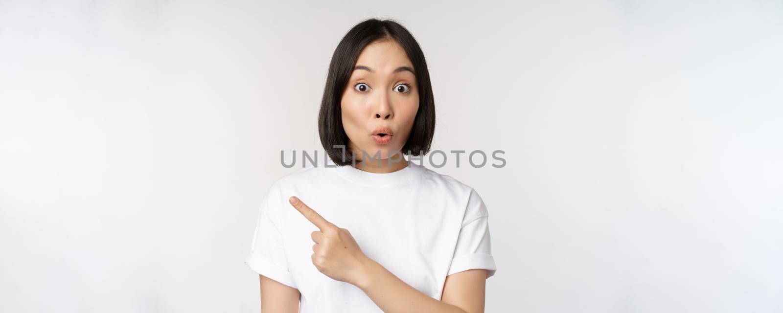 Close up of young japanese woman smiling, pointing finger left at copy space, showing announcement or advertisment banner, standing over white background.