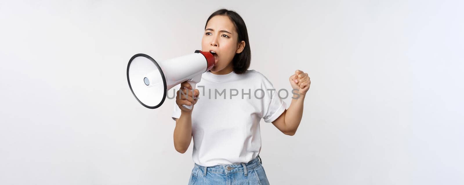 Asian girl shouting at megaphone, young activist protesting, using loud speakerphone, standing over white background. Copy space