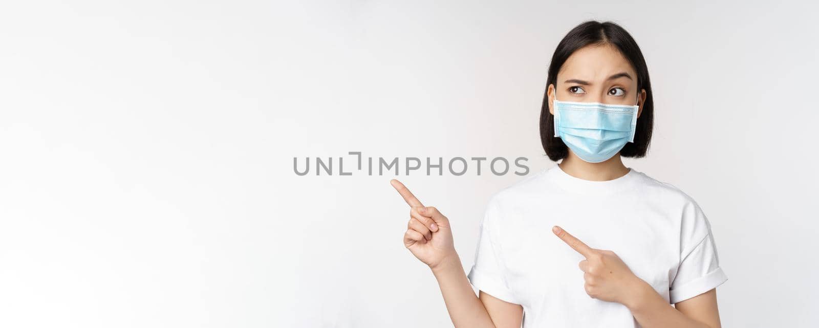 Image of asian woman in medical mask from covid, looking confused left, pointing at logo, showing advertisement, standing over white background.