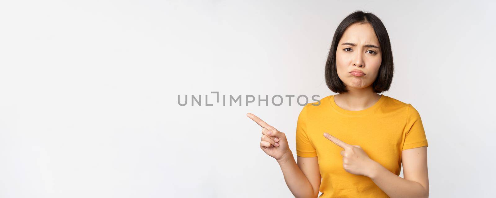 Portrait of disappointed brunette asian girl, pointing fingers left, grimacing upset, showing smth unpleasant, standing over white background by Benzoix