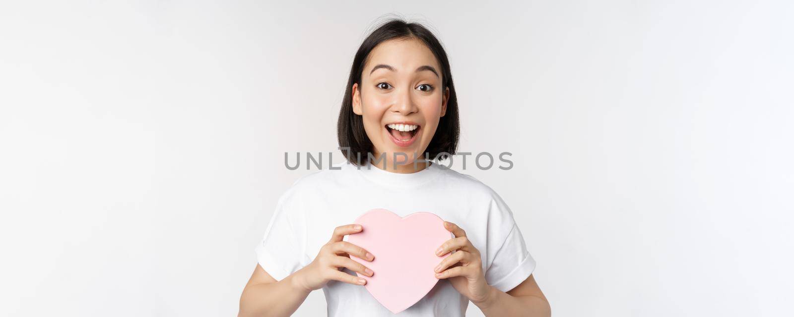 Valentines day. Happy asian girl receive romantic gift, holding heart shaped box and smiling excited, standing over white background by Benzoix