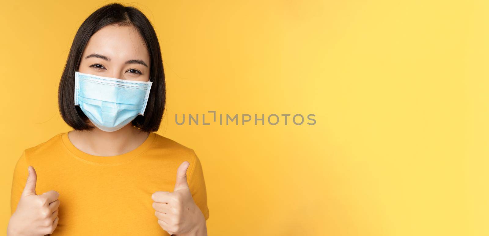 Cheerful korean woman in medical face mask, support people during pandemic, wear personal protective equipment from covid-19, showing thumbs up in approval, yellow background by Benzoix