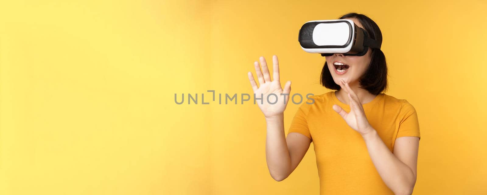 Girl in VR. Beautiful young asian woman, using virtual reality glasses and playing, chatting virtually, standing over yellow background by Benzoix