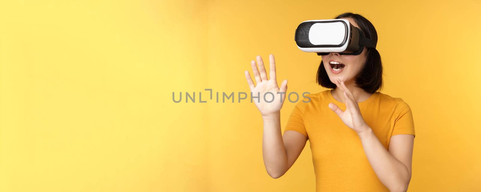 Girl in VR. Beautiful young asian woman, using virtual reality glasses and playing, chatting virtually, standing over yellow background.