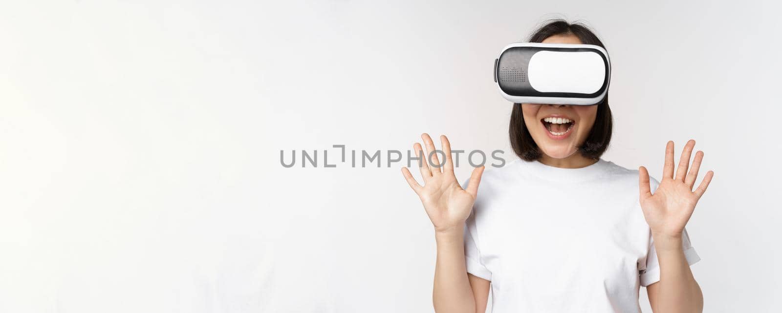 Happy asian woman using VR headset, waving raised hands and laughing, using virtual reality glasses, standing over white background.