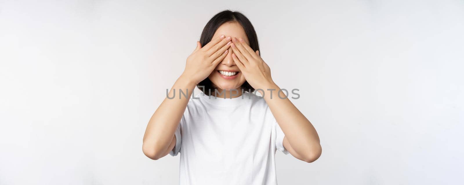 Portrait of asian woman covering eyes, waiting for surprise blindfolded, smiling happy, anticipating, standing against white background.