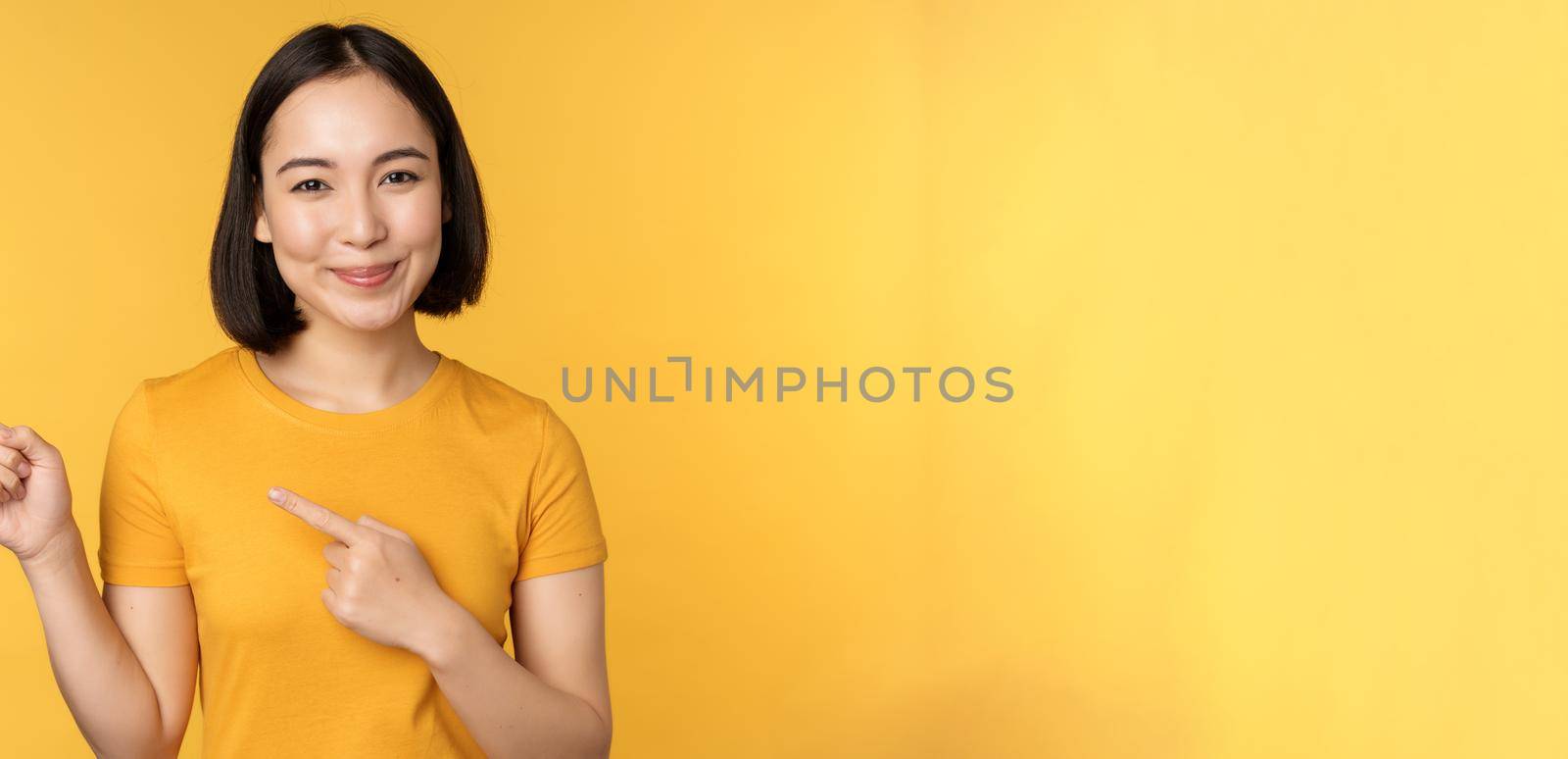 Smiling asian woman pointing fingers left, showing advertisement on empty copy space, standing over yellow background.