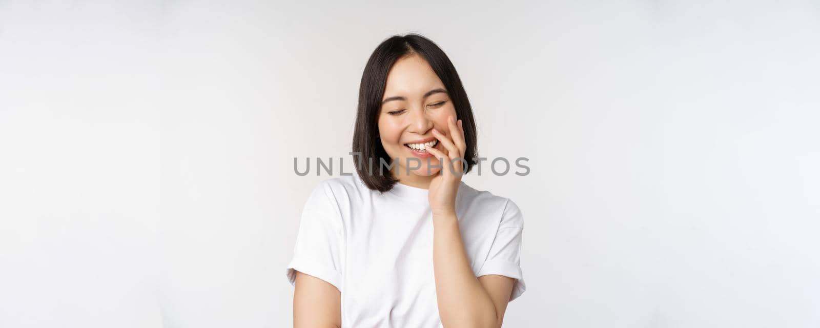 Portrait of young beautiful woman, korean girl laughing and smiling, looking coquettish, standing against white background.