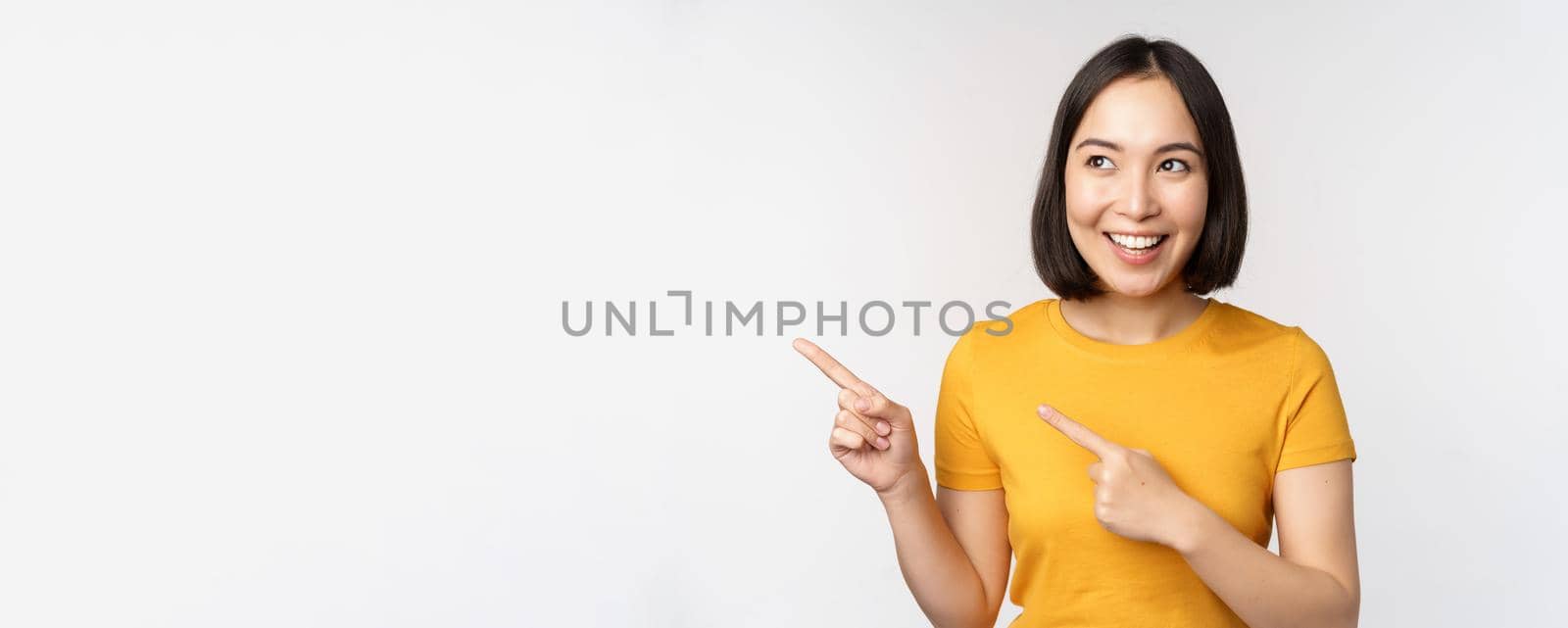 Portrait of smiling asian brunette girl in yellow tshirt, pointing fingers left, showing copy space, promo deal, demonstrating banner, standing over white background by Benzoix