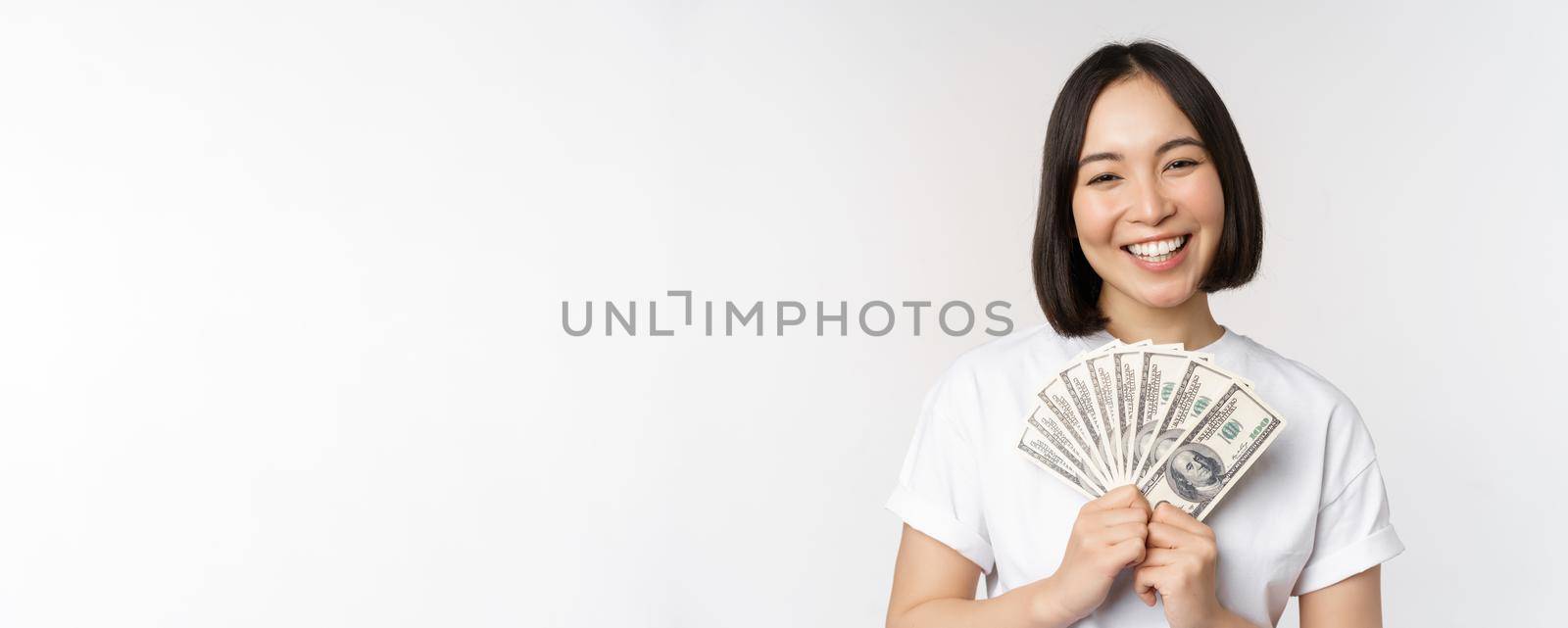 Portrait of smiling asian woman holding dollars money, concept of microcredit, finance and cash, standing over white background.