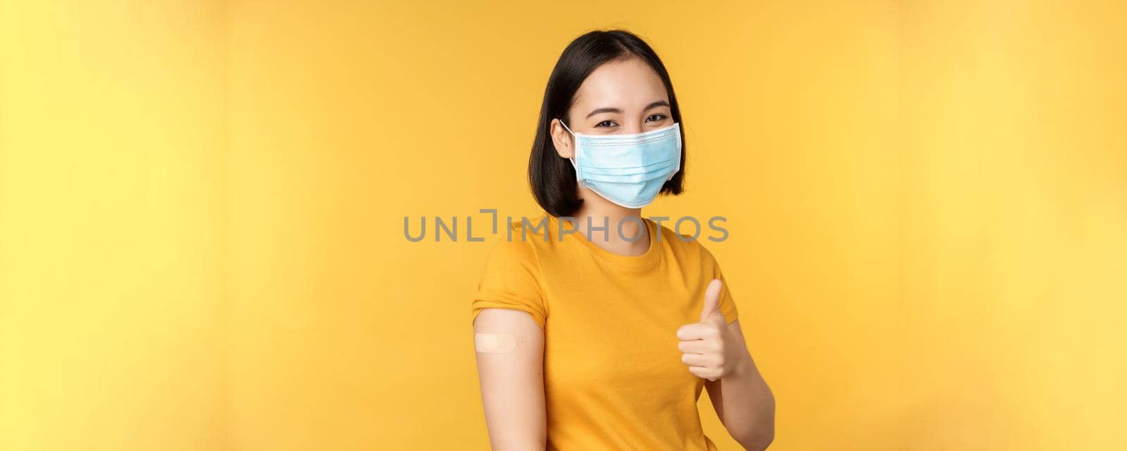 Vaccination from covid and health concept. Happy asian girl showing thumbs up, wearing medical mask, band aid on shoulder, got coronavirus vaccine shot, yellow background by Benzoix